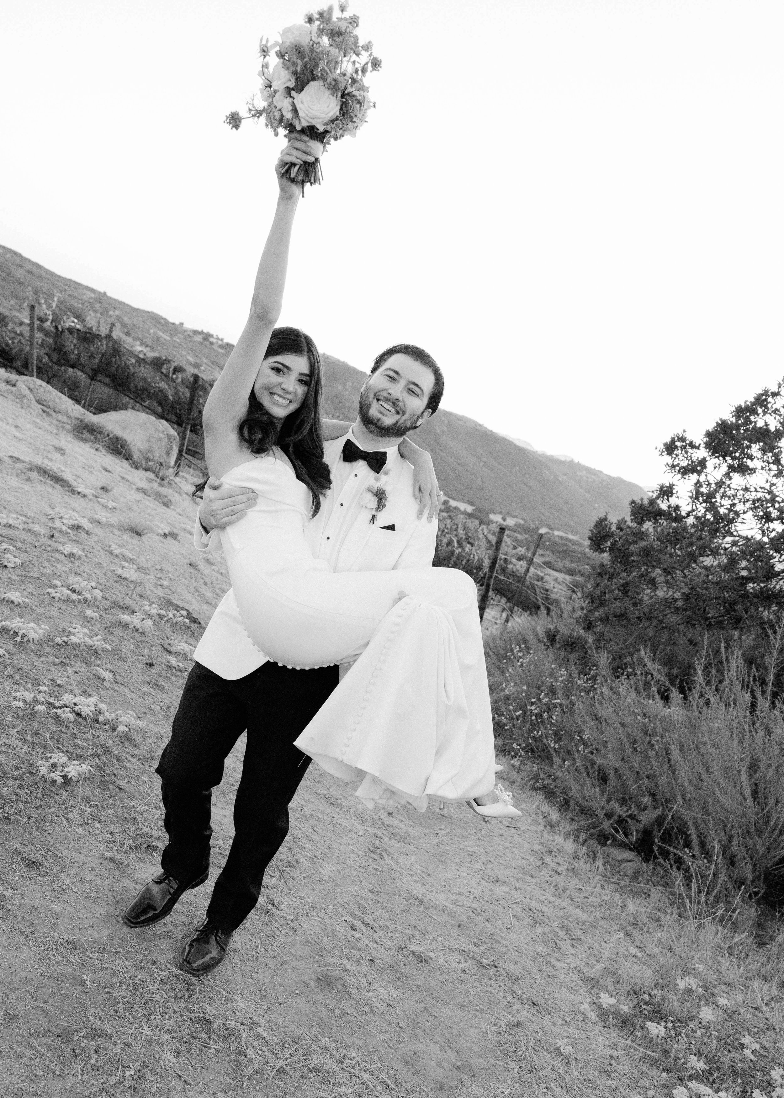 Joyful newlyweds celebrate on hillside path with raised bouquet in black and white.