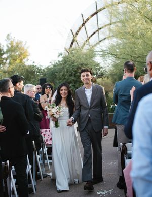 A couple walks down an outdoor aisle while guests look on at a garden wedding ceremony.