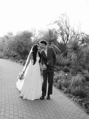 A romantic black and white wedding photo of a couple sharing a kiss on a brick pathway surrounded by desert plants.