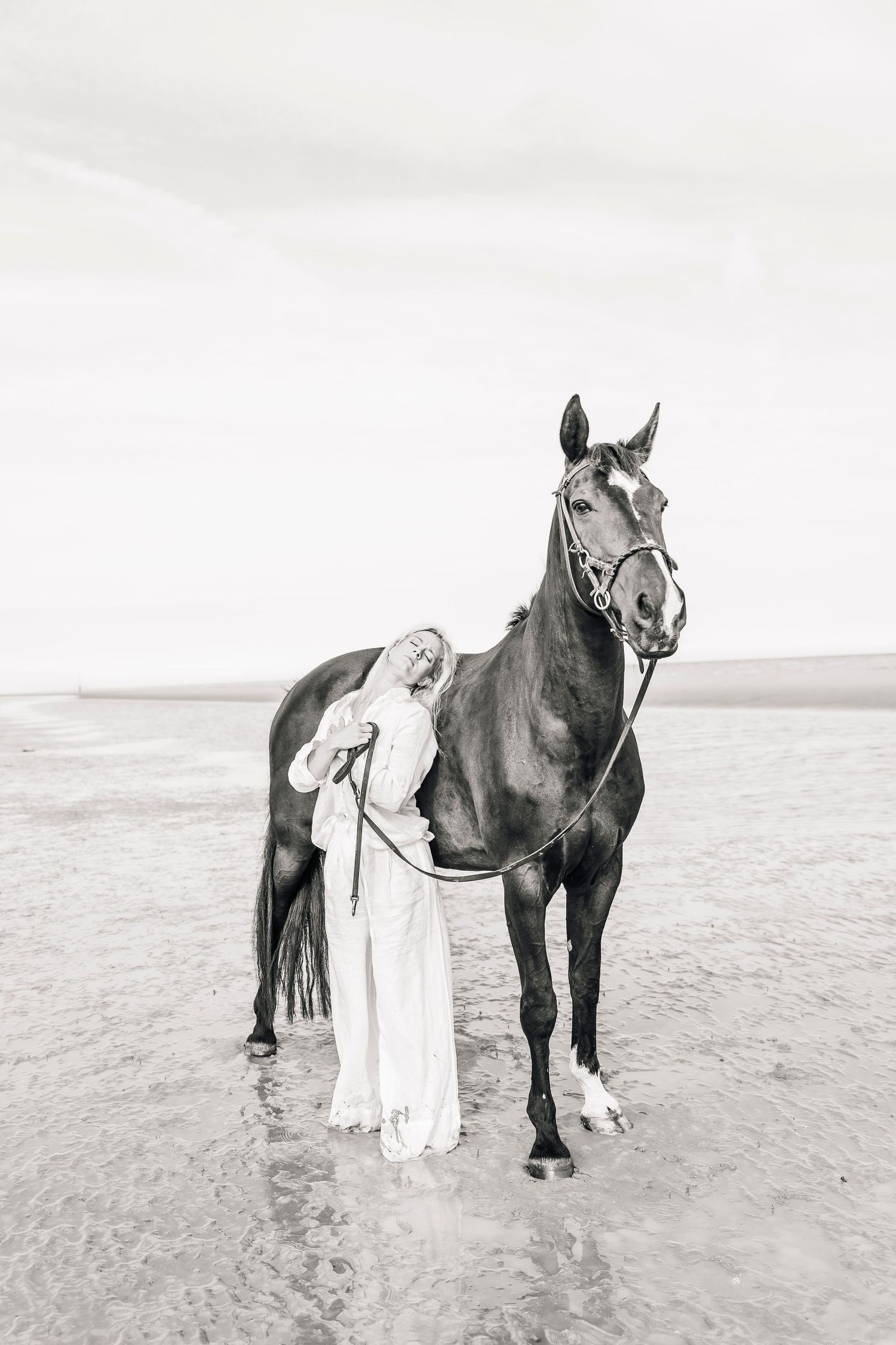 A majestic dark horse poses with someone in white on a misty beach in black and white.