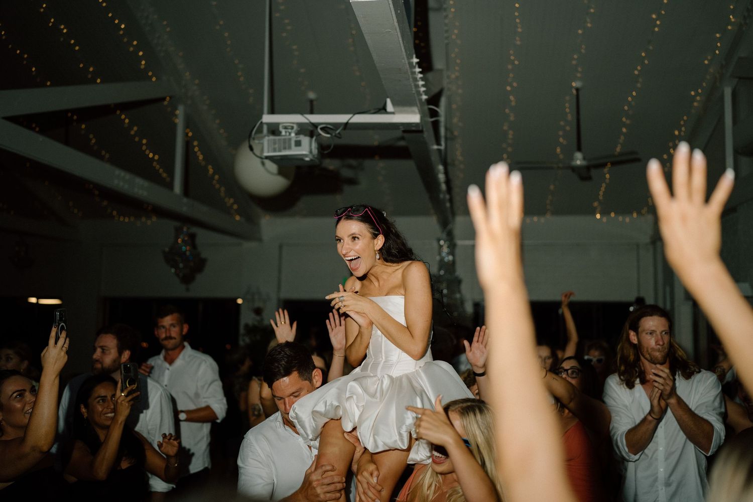 Person in a white dress being lifted up by a crowd at a lively indoor celebration party.