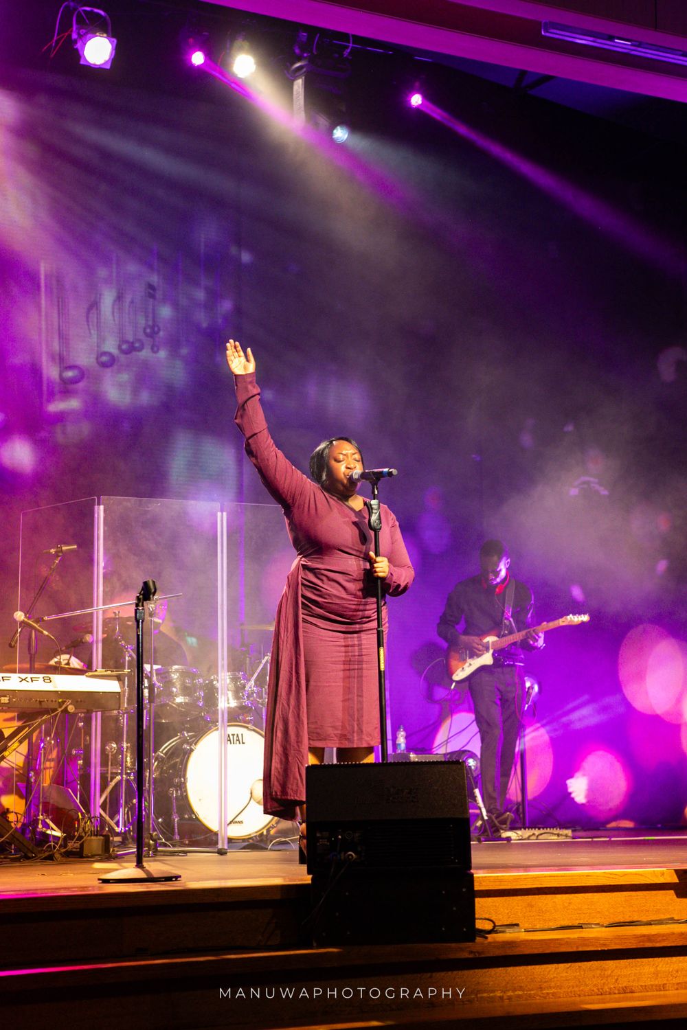 Concert performer in burgundy dress raises hand during emotional performance on stage with dramatic purple lighting.
