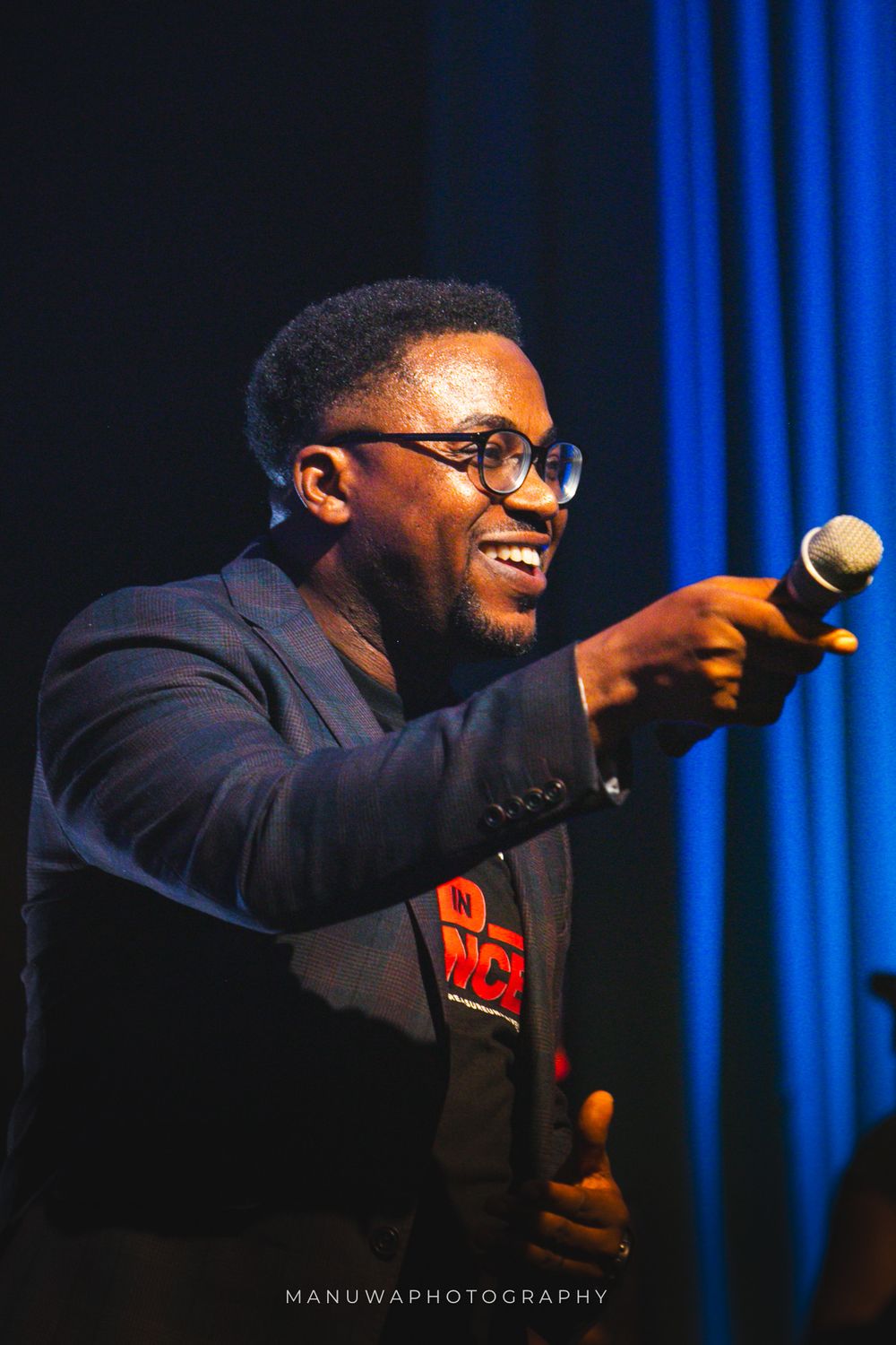 Performer in dark blazer smiles while gesturing with microphone during stage performance.