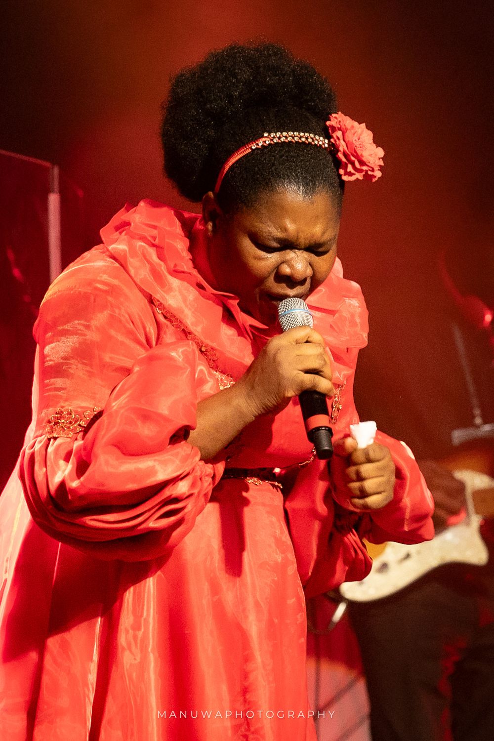 Singer in bright coral dress performs passionately on stage with dramatic red lighting.