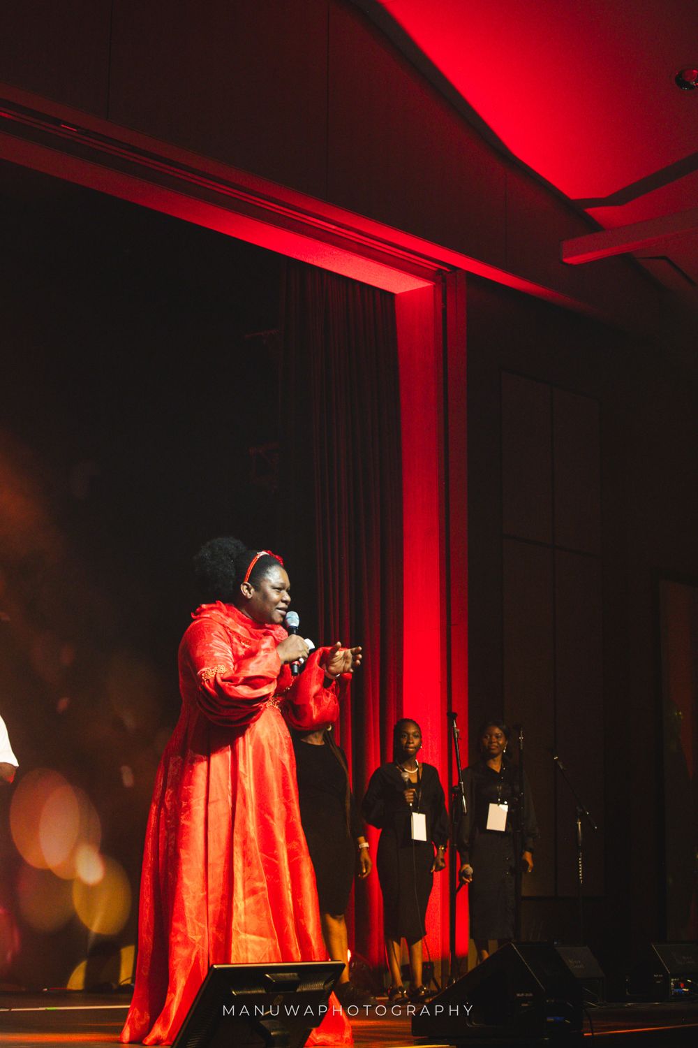 Performer in flowing red dress sings on dramatically lit stage with backup singers in background.