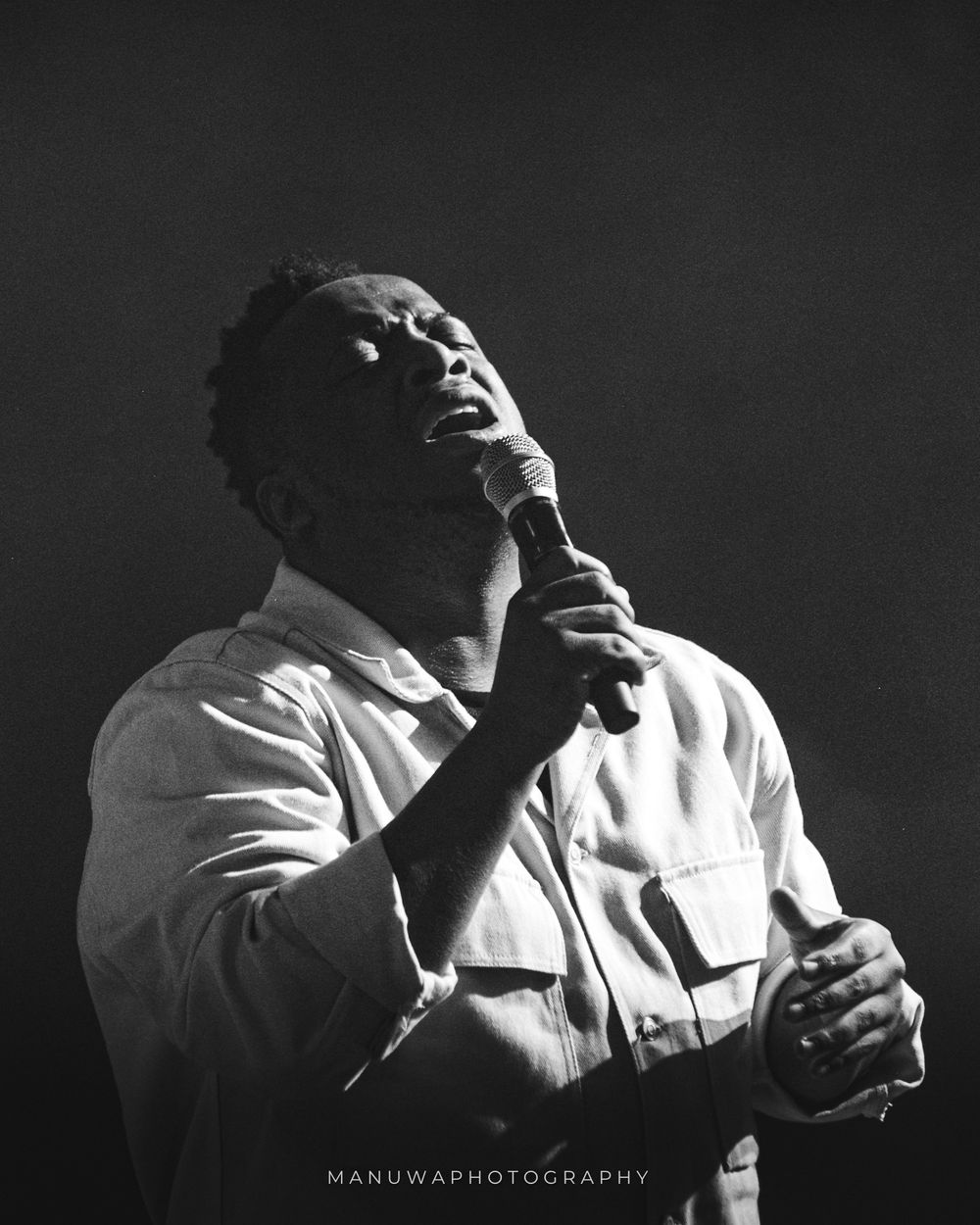 Black and white portrait of performer singing passionately into microphone on stage.