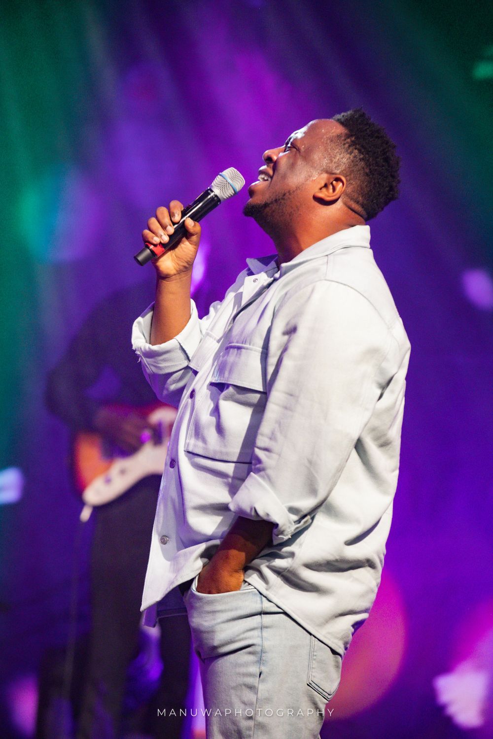 Singer in light colored shirt performs against vibrant purple and teal stage lighting.