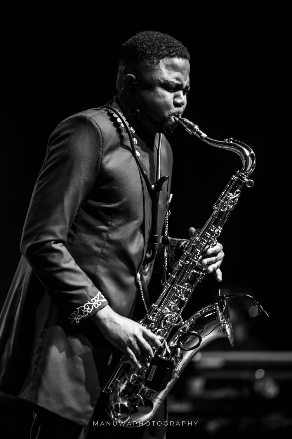 Saxophone player in formal attire performing on stage in black and white.