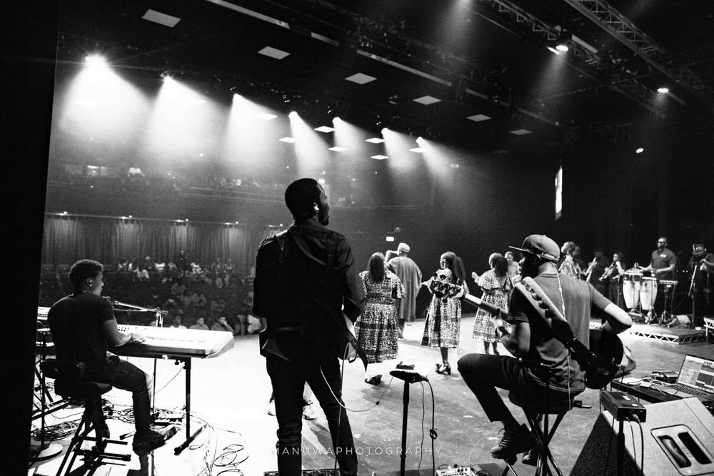Black and white concert photo showing silhouettes of performers and band on dimly lit stage.