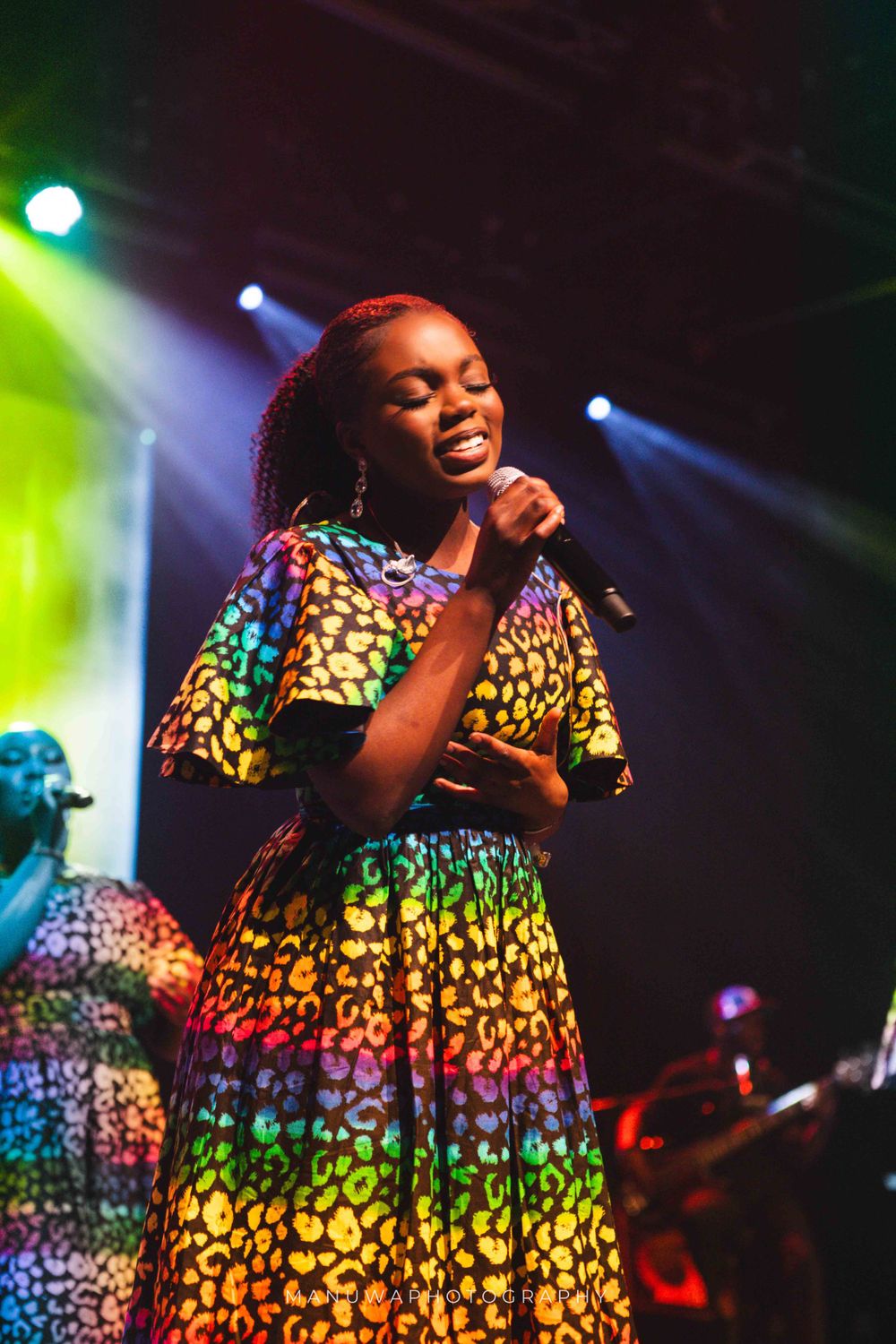 Singer performing in vibrant multi-colored African print dress under stage lights.