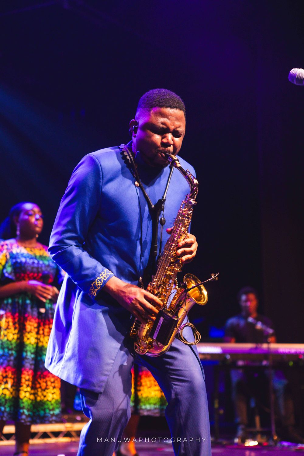 Saxophone player in blue suit performing passionately with backup singers in colorful dresses.