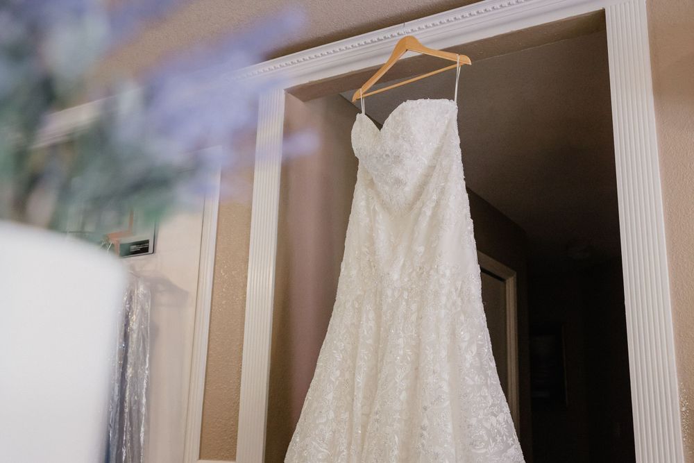 A white lace wedding dress hanging elegantly on a hanger against a beige wall.