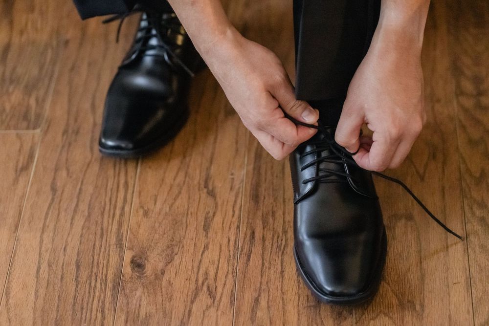 Hands tying the laces of formal black leather dress shoes on wooden flooring.