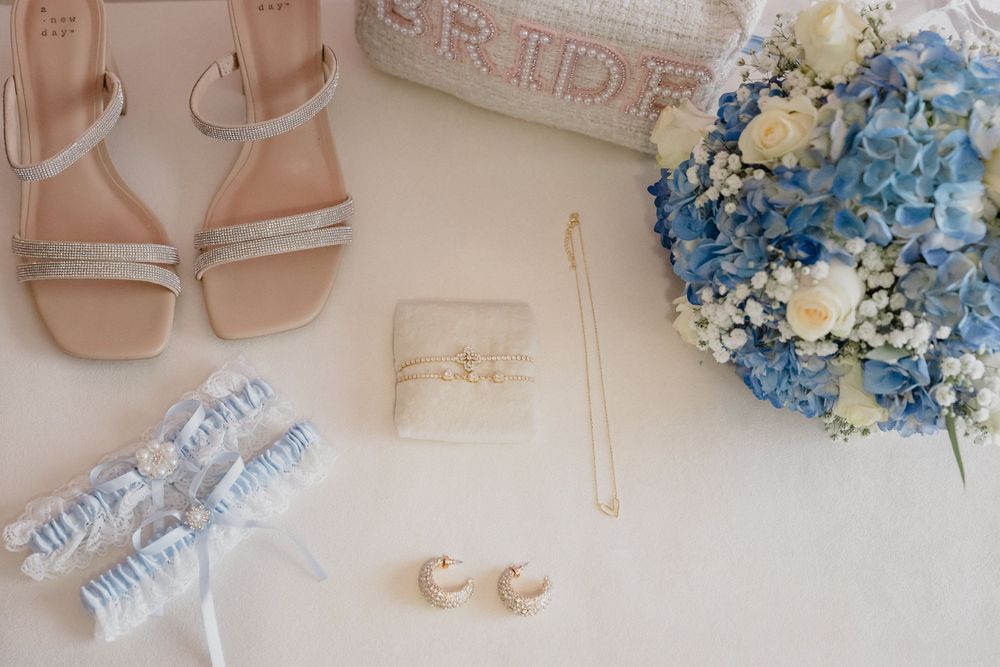 Wedding accessories including sandals, garter, blue bouquet and jewelry on cream surface.