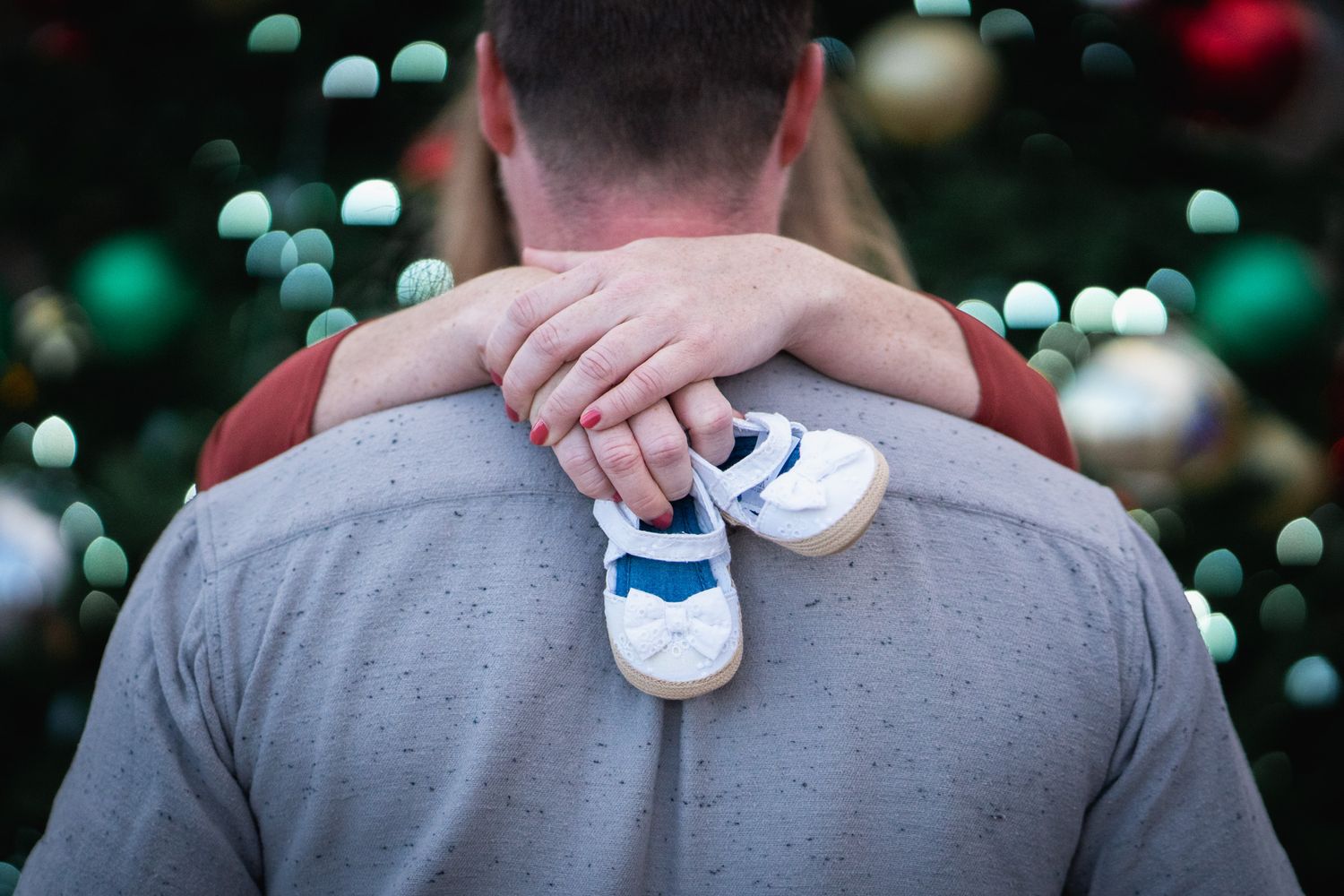 Close-up of tiny white baby shoes held against grey shirt with Christmas lights in background.