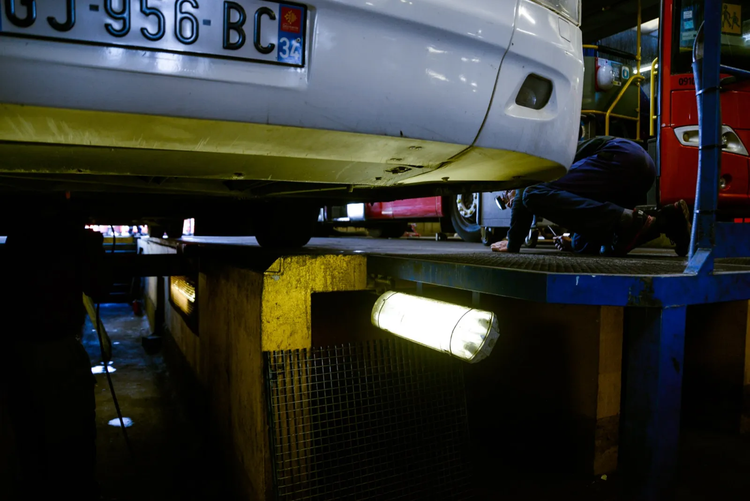 Bas de caisse d’un véhicule en réparation, éclairage jaune sous le châssis et outillage visible autour.
