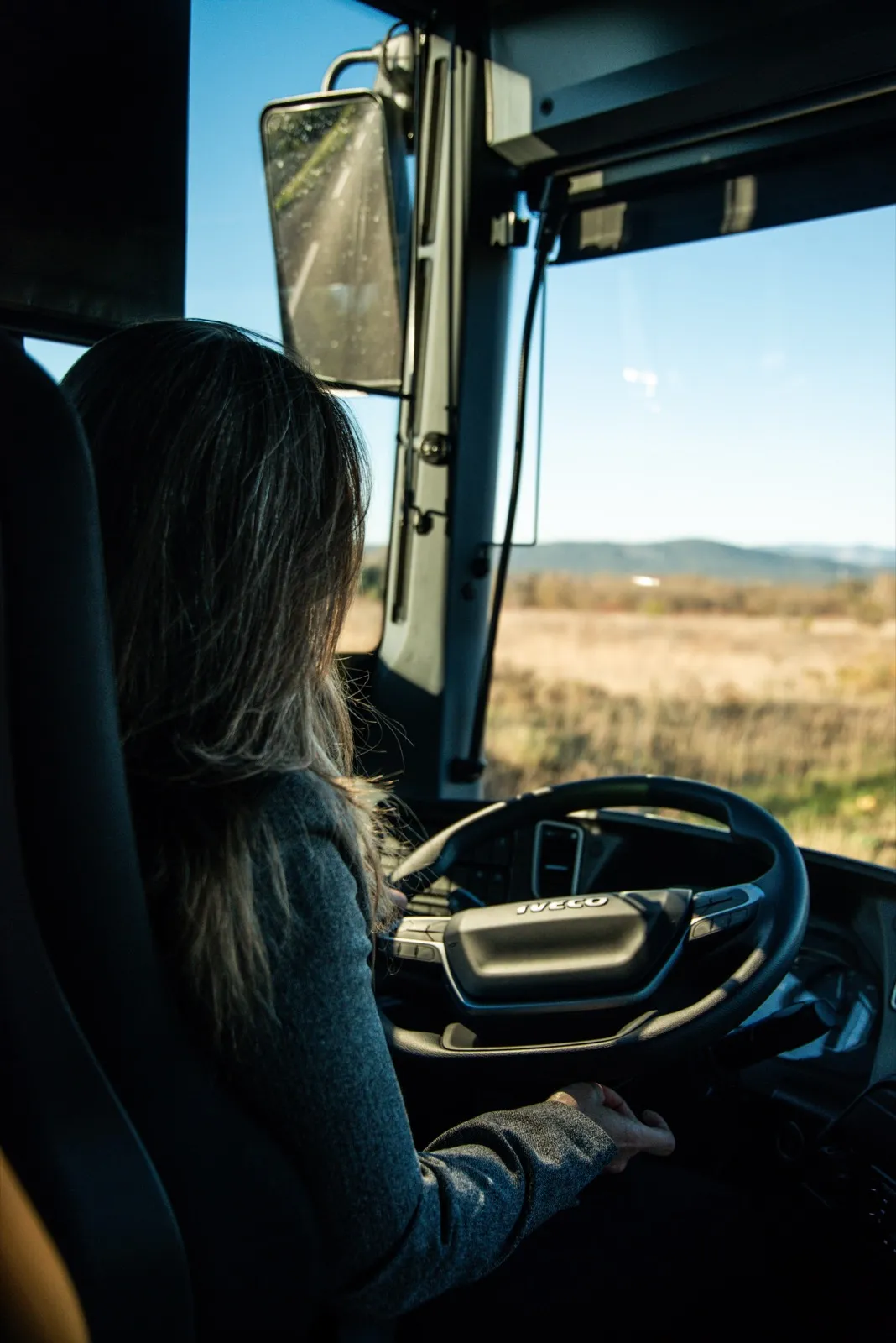 Femme au volant d’un bus, concentrée sur la route à travers la vitre baignée de lumière dorée.