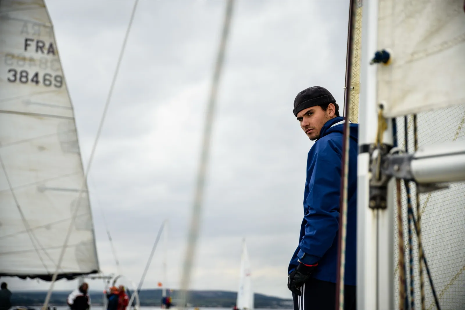 Jeune homme en ciré bleu debout sur un voilier, regard tourné vers l’horizon, entouré de voiles blanches.