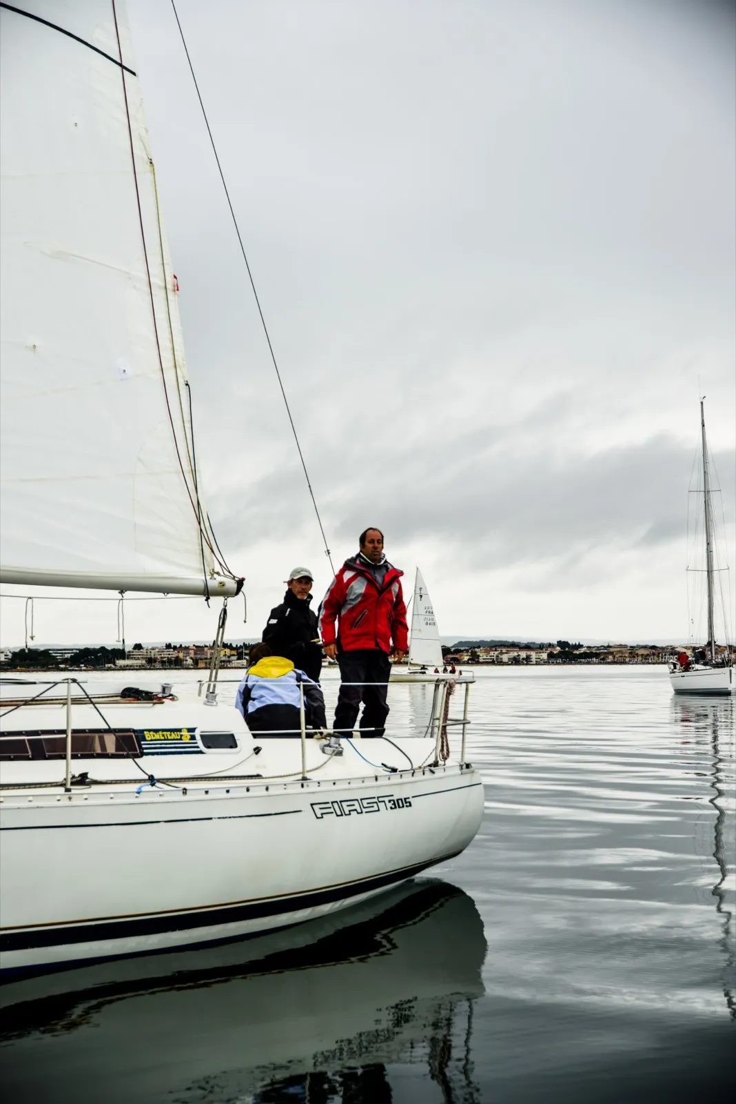 Deux personnes en ciré rouge à l’arrière d’un voilier, vues depuis un autre bateau dans un port calme.