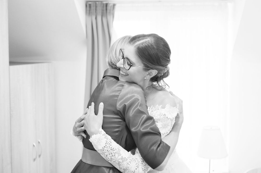 Black and white photo of a bride in lace dress warmly embracing someone in a bright room.