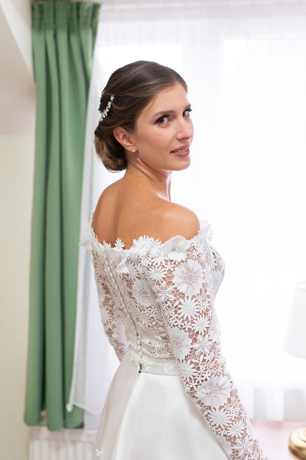 Beautiful bride in off-shoulder lace wedding dress glancing back over her shoulder indoors.