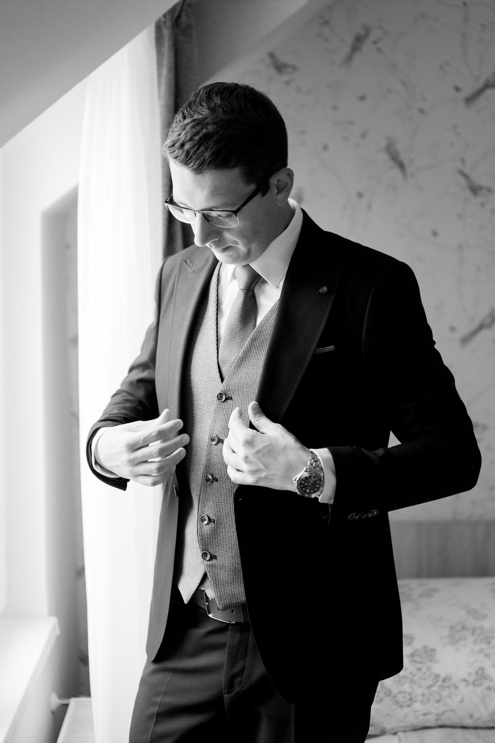 Black and white portrait of dapper groom adjusting his suit jacket by a bright window.