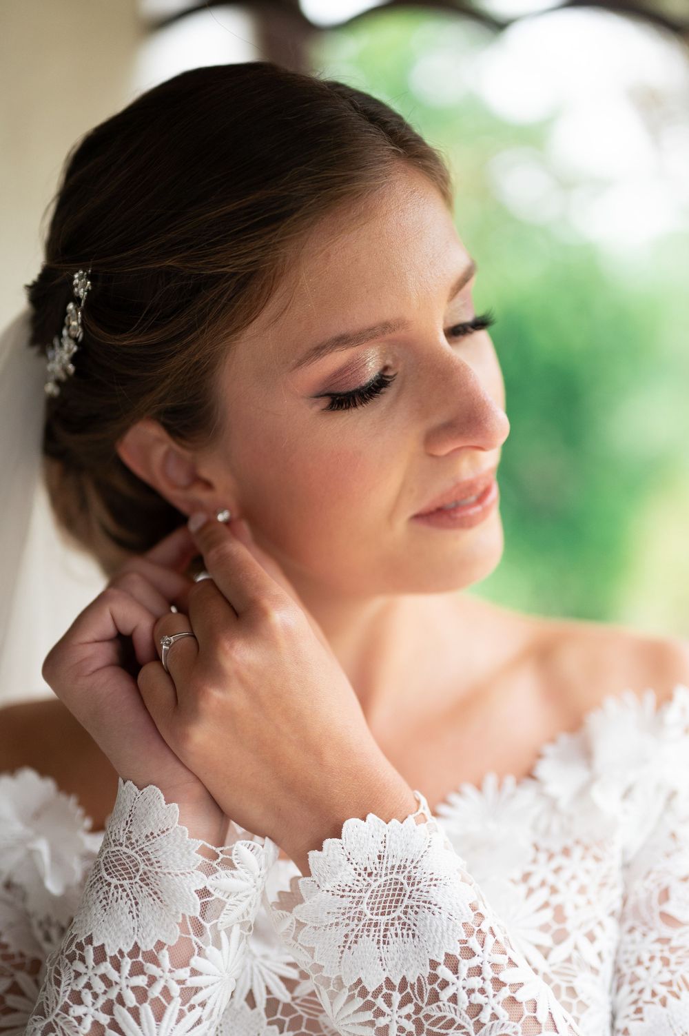 Bride in lace off-shoulder gown adjusting pearl earring with elegant updo and floral hair accessory.