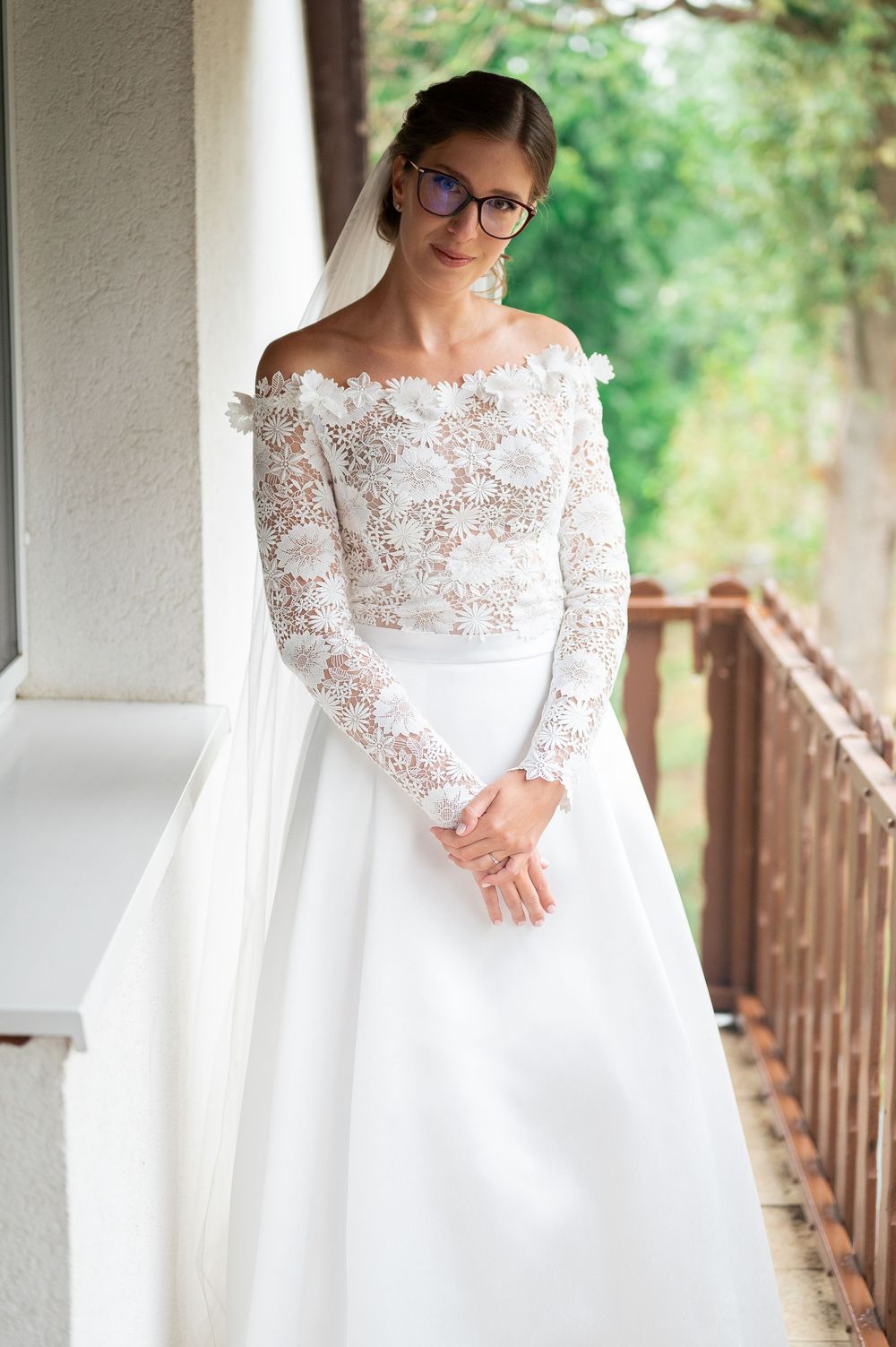 Elegant bride in off-shoulder lace wedding gown and veil posing on outdoor balcony.