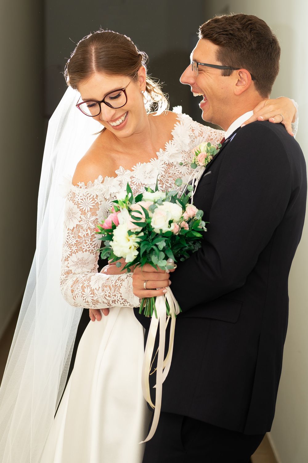 Laughing bride and groom embracing while bride holds white and pink wedding bouquet indoors.