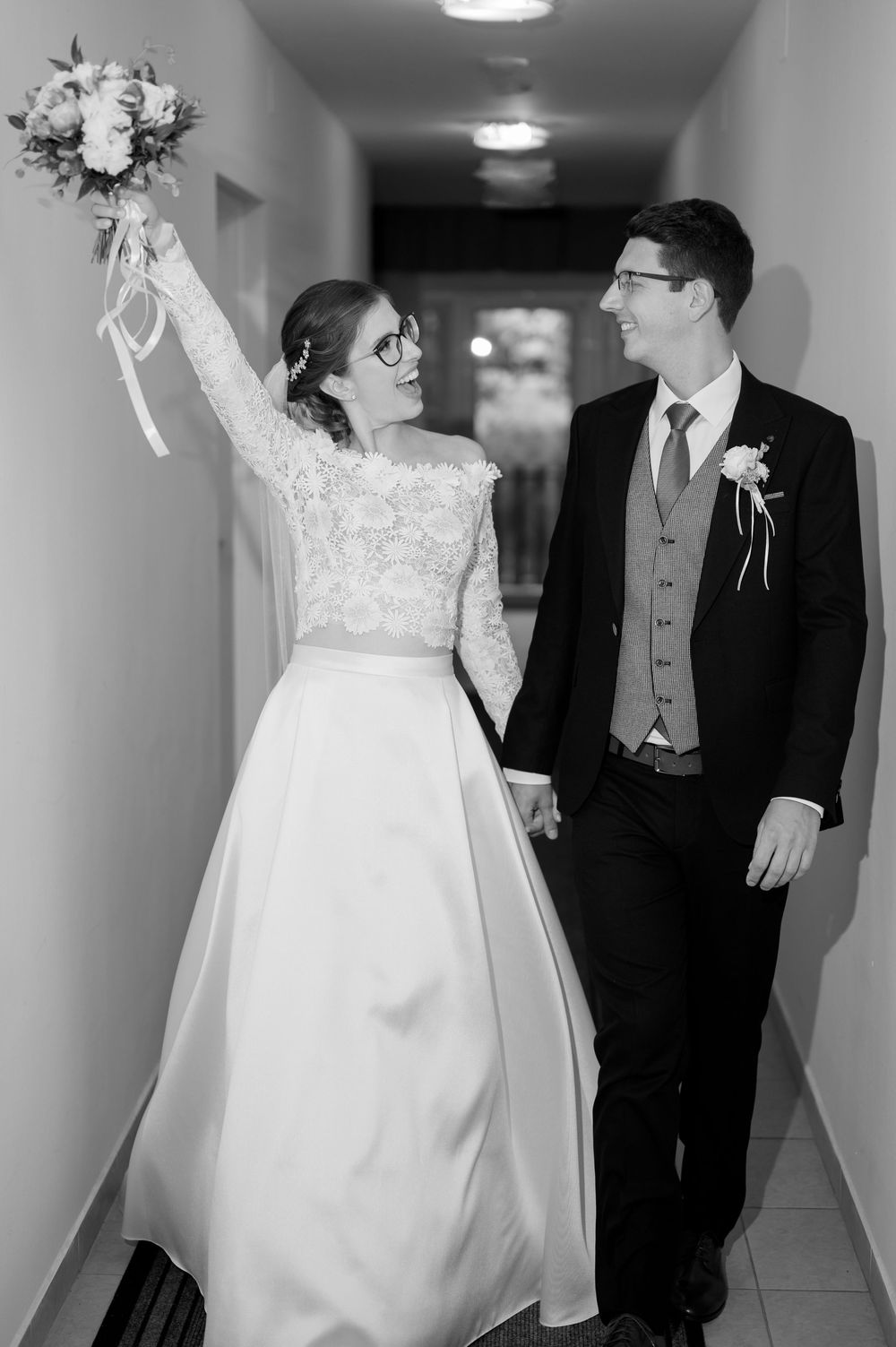 Black and white photo of joyful bride raising bouquet while holding grooms hand in hallway.