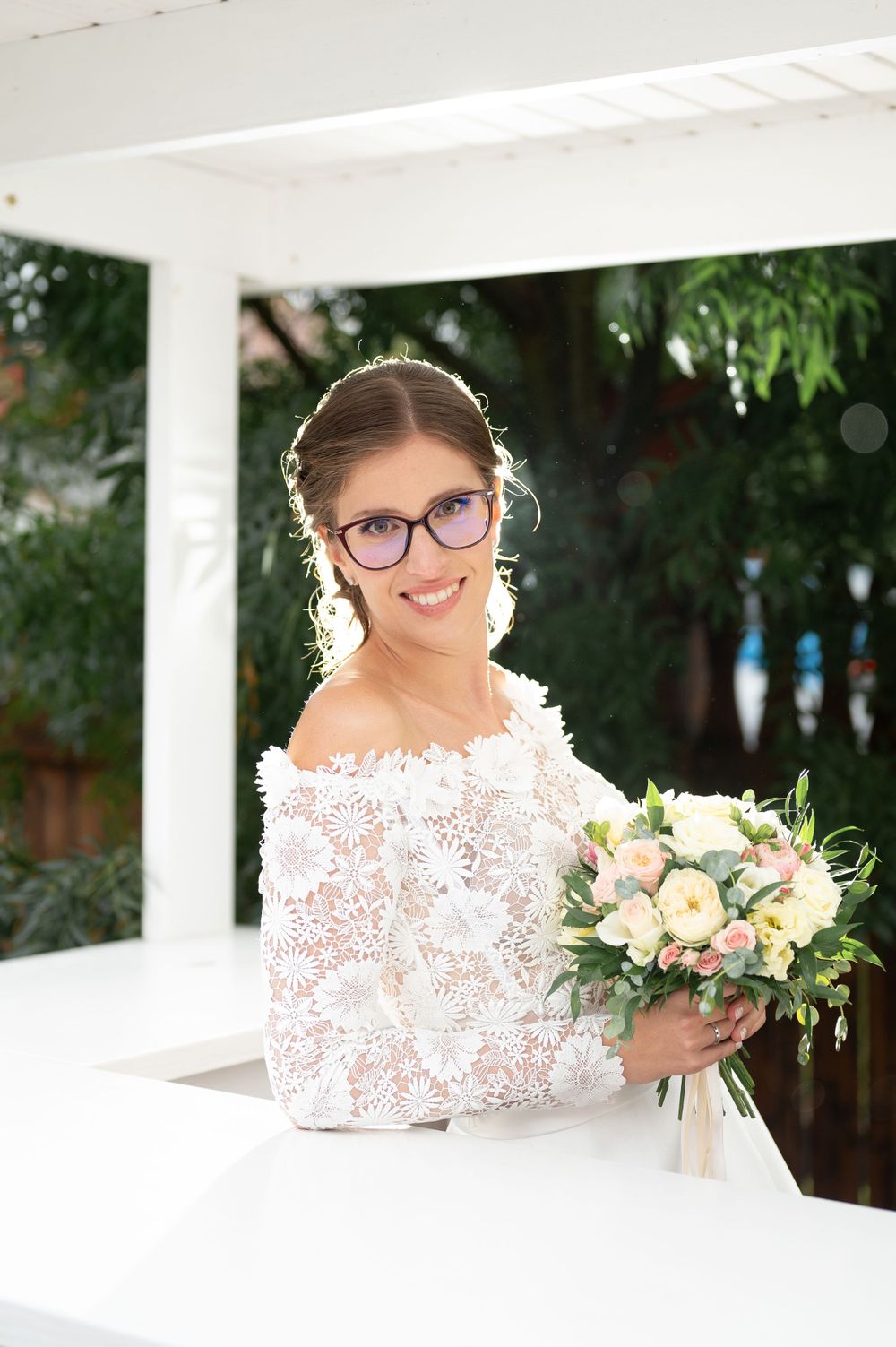 Smiling bride wearing glasses and white lace off-shoulder dress holding a pastel bouquet in a gazebo.