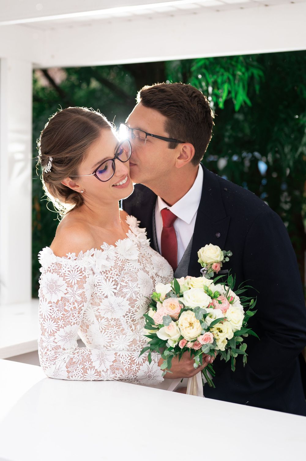 Groom kissing smiling bride on cheek as she holds pastel bouquet in outdoor wedding gazebo.