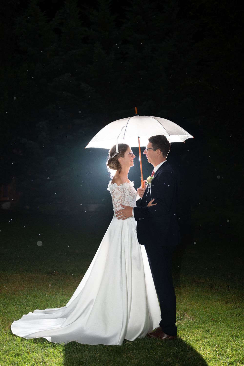 Bride and groom sharing a romantic moment under a white umbrella on a rainy wedding night.