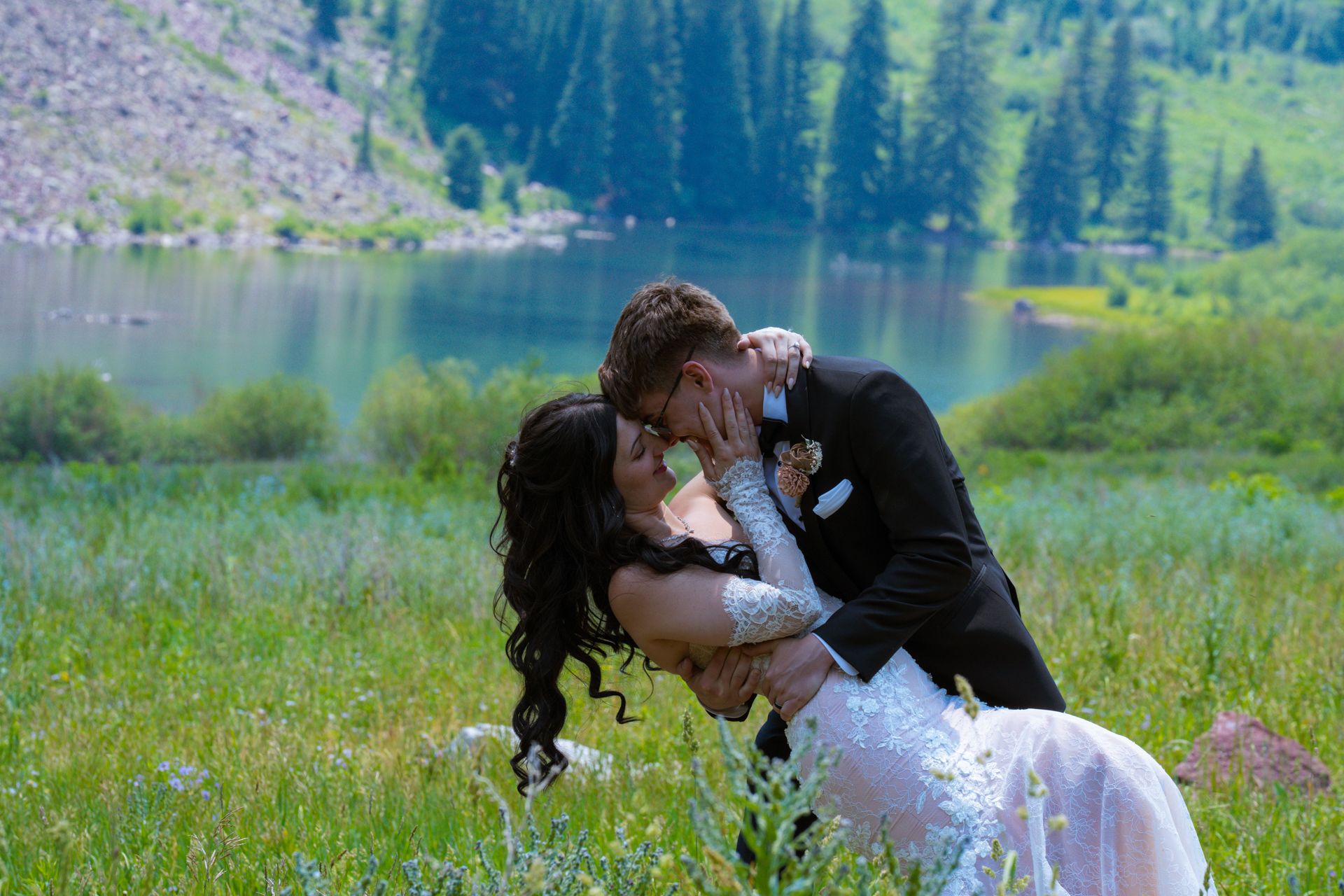A couple shares a romantic kiss while dipping in a lush green meadow beside a serene mountain lake.