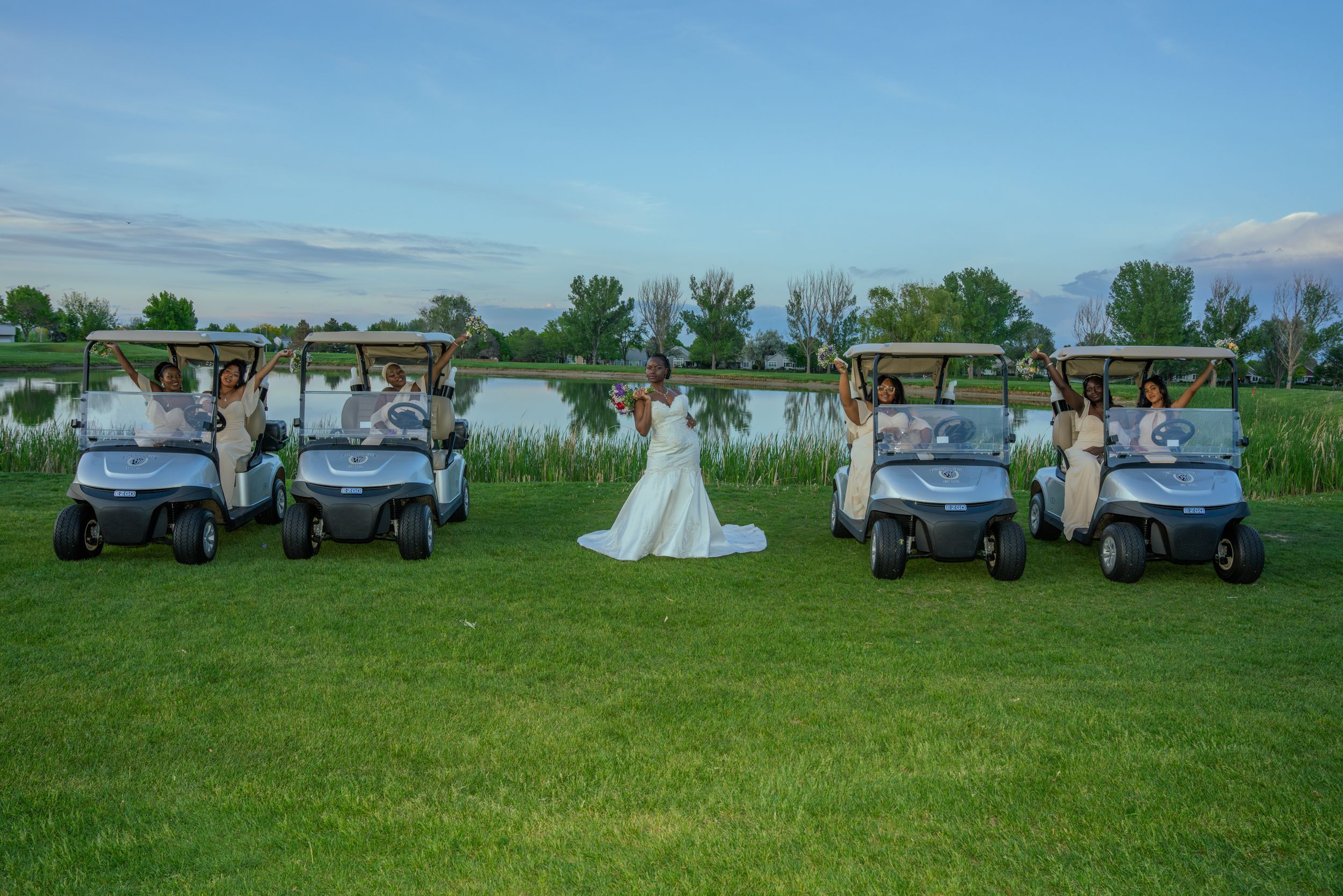 Wedding party poses with vintage cars in a green field during a sunny outdoor celebration.