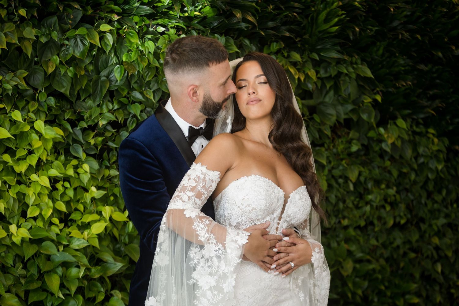 A romantic wedding portrait of a couple embracing against a lush green foliage wall outdoors.