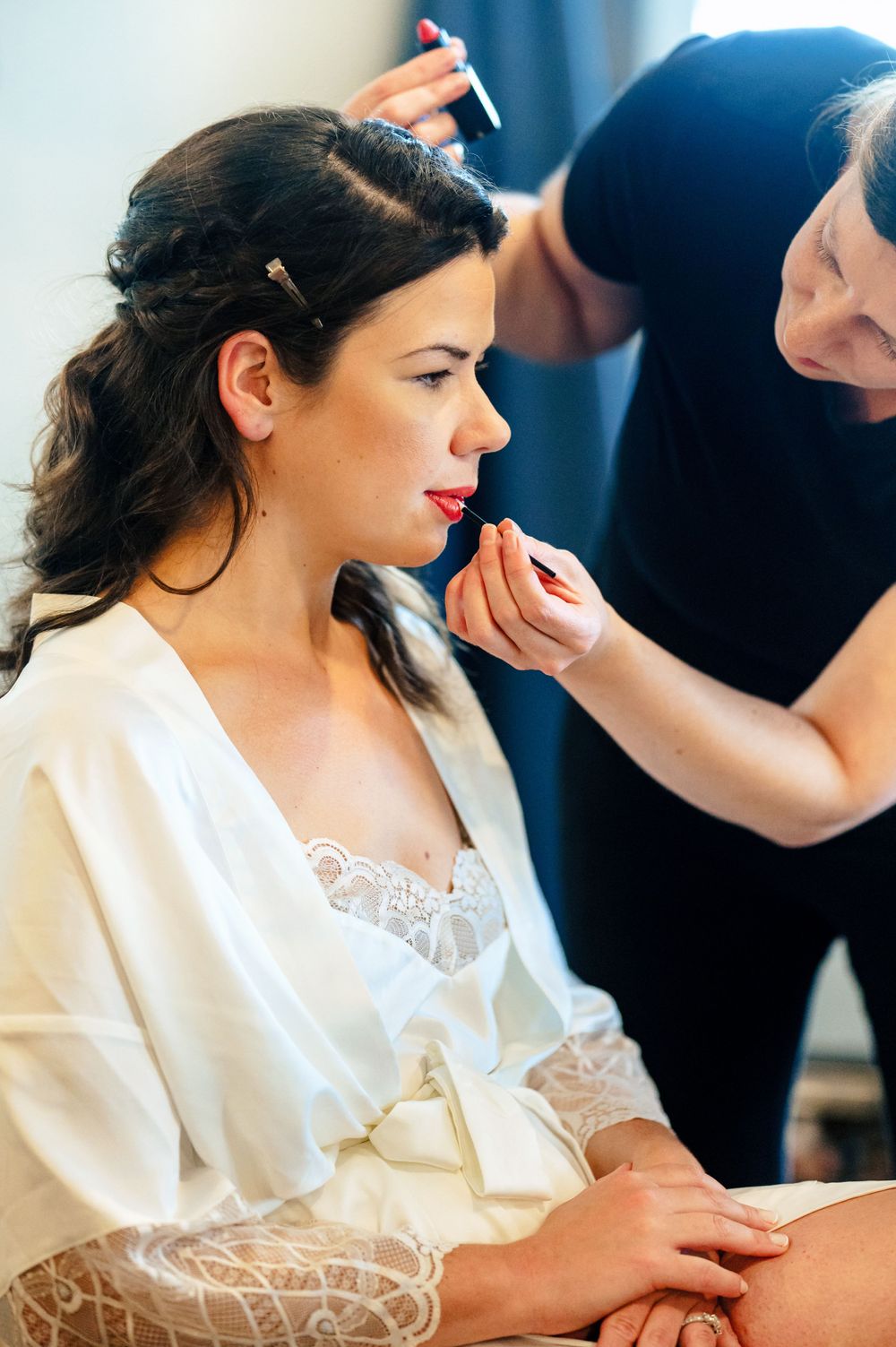 Makeup artist applies red lipstick to a dark-haired bride in a white lace robe.