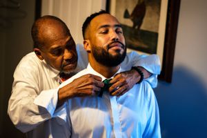 Older man helps young groom adjust his tie while getting ready for wedding.