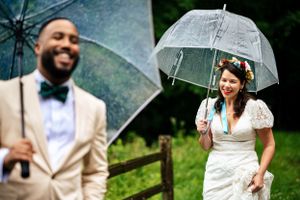 Smiling groom in tan suit holds clear umbrella as bride approaches in white dress.