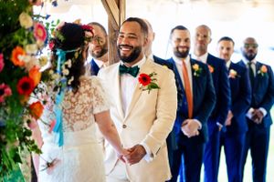 Groom in white tuxedo smiles at bride with groomsmen in navy suits standing behind them.