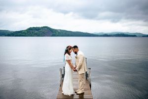 Bride and groom hold hands on a wooden dock with a scenic lake and mountains behind them.