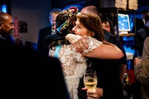 Bride in lace dress shares an emotional hug with a woman at an indoor wedding reception.