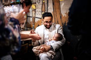 Groom in white suit holds a sleeping newborn baby at a rustic wedding venue.