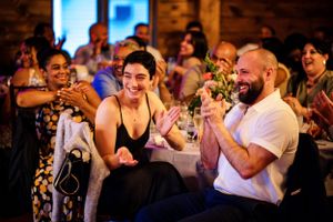 Wedding guests laughing and clapping joyfully during reception celebration indoors.