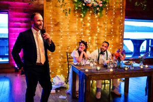 Man delivers a wedding speech as bride and groom smile at a rustic barn reception.