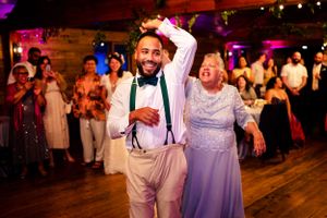 Groom dancing joyfully with older woman at wedding reception in rustic barn venue.