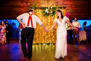 Bride and groom hold hands dancing before floral fairy light backdrop at wedding.