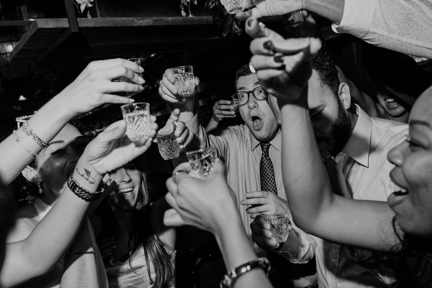 Black and white photo of excited party guests raising drinks in a toast at a lively celebration event.