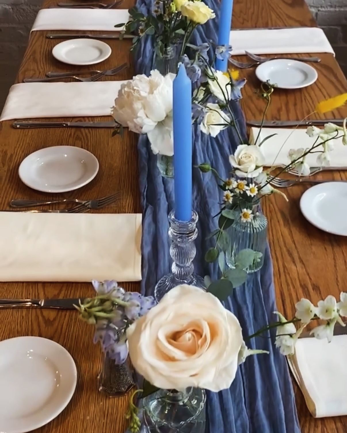Elegant table setting with white plates, cream roses, and blue taper candles on a wooden surface with navy blue table runner.