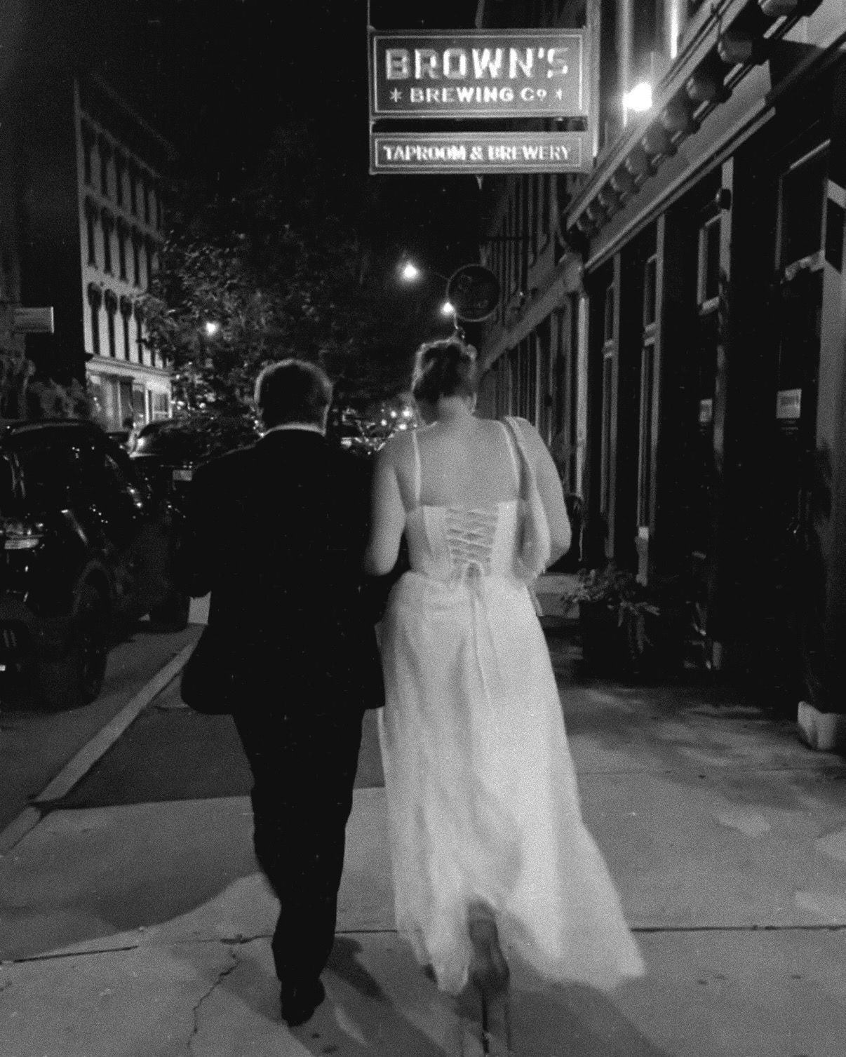 Couple in wedding attire strolling under a brown's shop sign at night in urban setting.